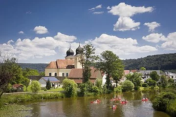 Auf der Altmühl paddeln fünf rote Kanus an Kottingwörth mit ihrer Kirche und deren zwei Kirchtürmen vorbei.