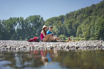 Ein Pärchen mit Hüten sitzt am Kiesstrand des Kloster Weltenburg. Vor ihnen fließt die Donau vorbei und im Hintergrund ist der Wald gegenüber zu sehen.