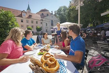 Im Biergarten des Klosters ist der Blick auf einen Biertisch gerichtet. Er ist mit weißer Tischdecke sowie einer kleineren weiß-blaukarierten Tischdecke gedeckt. Darauf ist im Vordergrund ein Korb mit Laugenbrezen gefüllt. An dem leeren Platz wurde der dunkelrote Fahrradhelm an die Stuhllehne gehängt. Es sitzen drei Frauen und zwei Männer an dem gedeckten Tisch, in der Mitte steht eine Brotzeitplatte. Im Hintergrund ist das Gebäude des Klosters und der Blick in den gut gefüllten Biergarten im Schatten zu sehen.