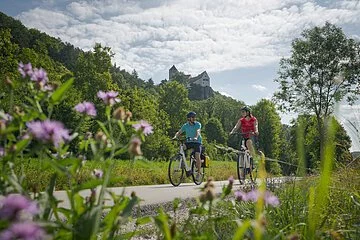 Zwei Radler, eine Frau mit weißem Helm und roten Shirt und ein Mann, neben ihr, ebenfalls mit Helm und blauen Shirt sind auf dem Radweg unterwegs. Die Zwei radeln und hinter ihnen ragt die Burg Prunn, die umgeben von Wäldern auf einem Felsen. Der Himmel scheint blau, nur ein paar Wolken verirren sich.