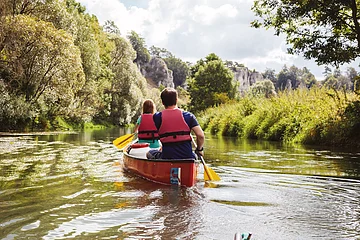 Ein Mann und eine Frau mit dem Rücken zu uns, sitzen in einem roten Kanu auf der Altmühl. Die gelben Paddel erzeugen leichte Wellen auf der Wasseroberfläche. im Hintergrund ist das grüne Ufer mit prächtigen Bäumen zu sehen und rechts blitzt der blaue Himmel durch.